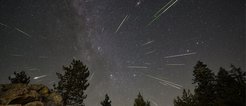 This image shows the Perseid meteor shower in Sequoia National Park, California, 2023. Streaks of shooting stars with bright, fiery trails cut through the dark night sky. Silhouettes of tall tree tops frame the scene, with radiant lines of meteors in the background.