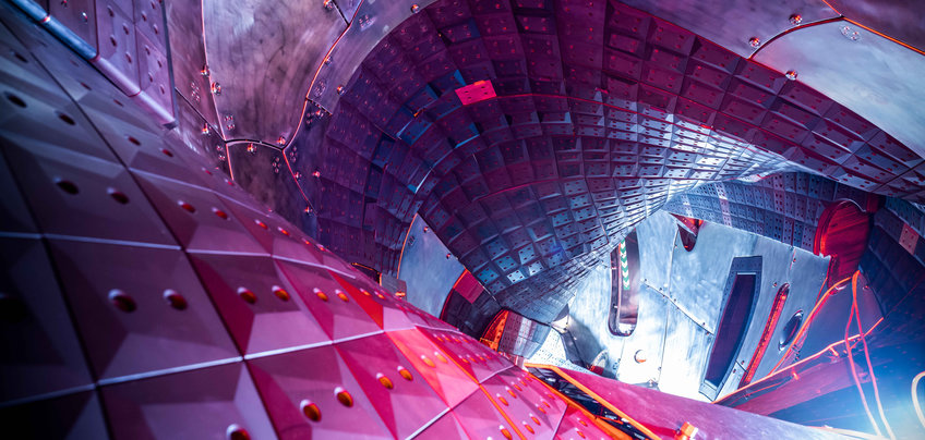 Ingenious twists and turns: The plasma chamber of Wendelstein 7-X (a German thermonuclear fusion experiment) is constructed like a twisted pretzel. The wall is lined with particularly robust graphite tiles. The interior of a futuristic, metallic structure featuring curved surfaces with geometric panels and bolts, illuminated by vibrant red and blue lighting, suggesting advanced technology.